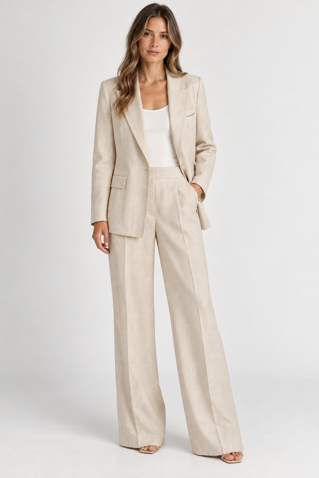 A woman modeling a light beige linen two-piece suit with a blazer and wide-leg trousers, styled over a white tank top and neutral sandals, against a plain light gray studio background.