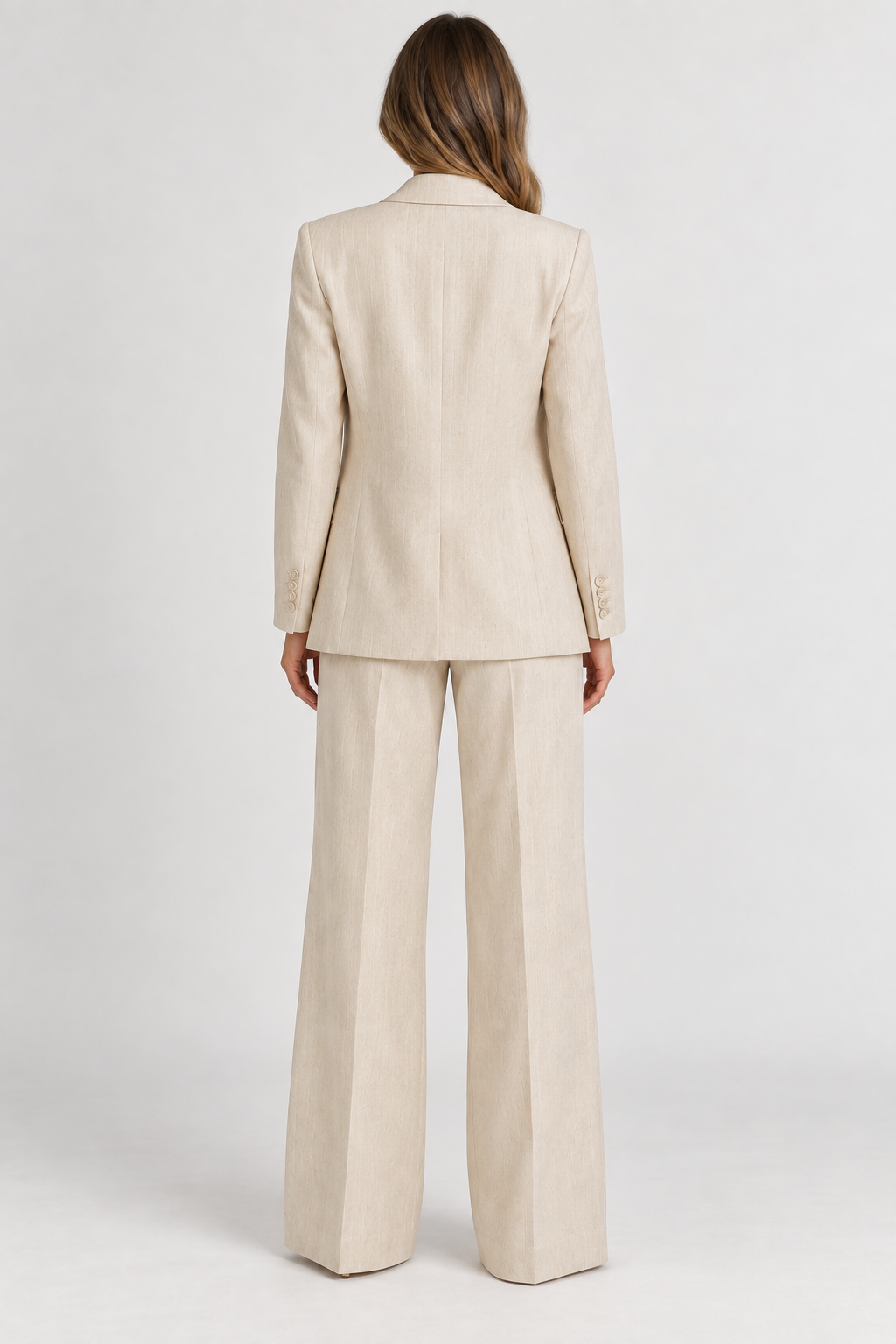 A woman viewed from the back wearing a light beige tailored suit with a blazer and wide-leg trousers, photographed against a clean light gray studio background.