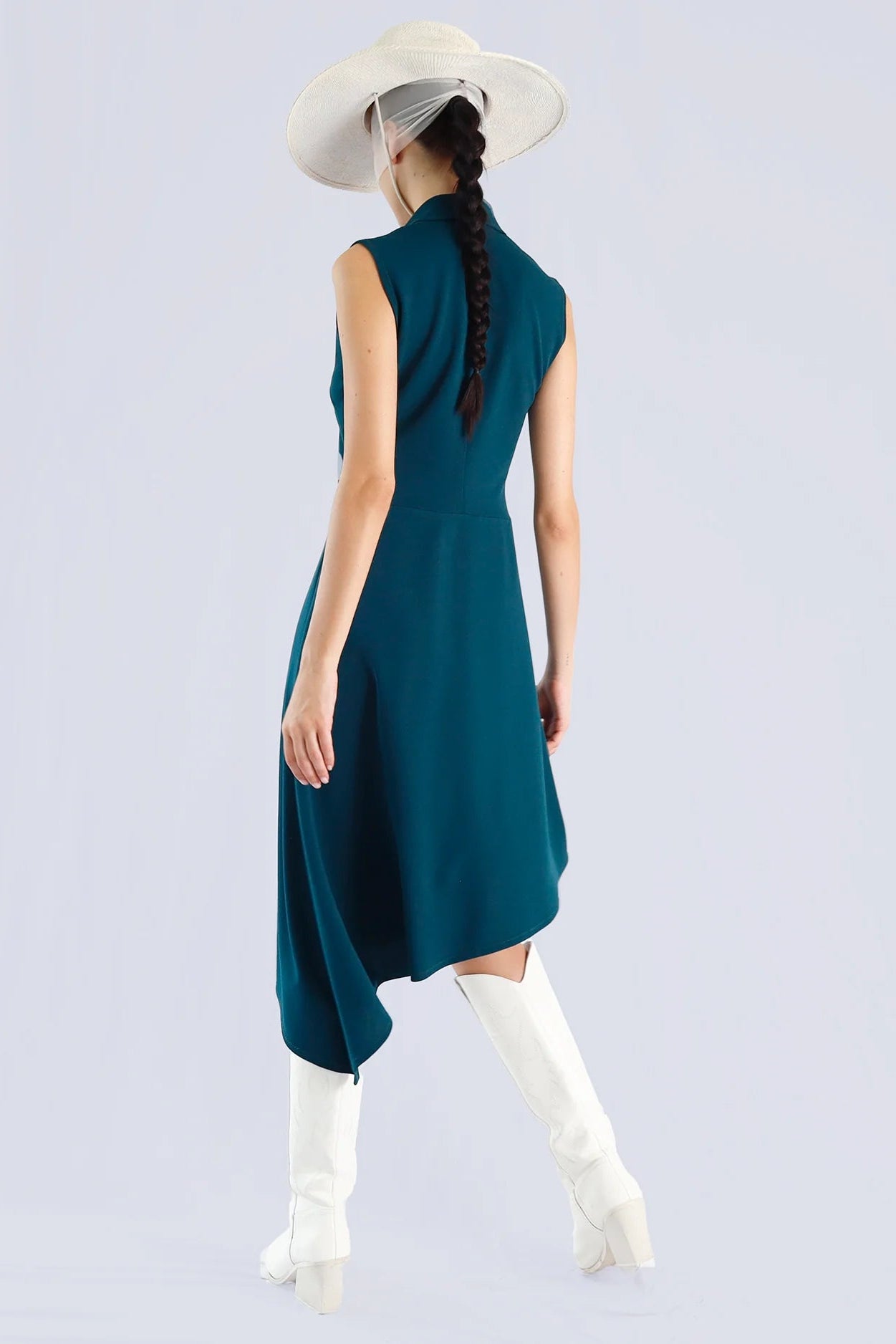 Back of a woman wearing a teal sleeveless dress with white boots and a wide-brimmed hat on a light gray background.