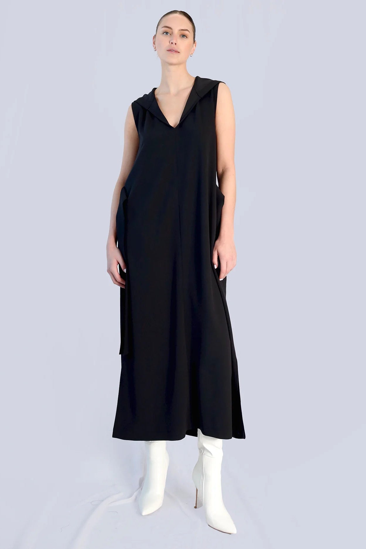 Woman wearing a long black dress, with heeled boots, on a grey background.