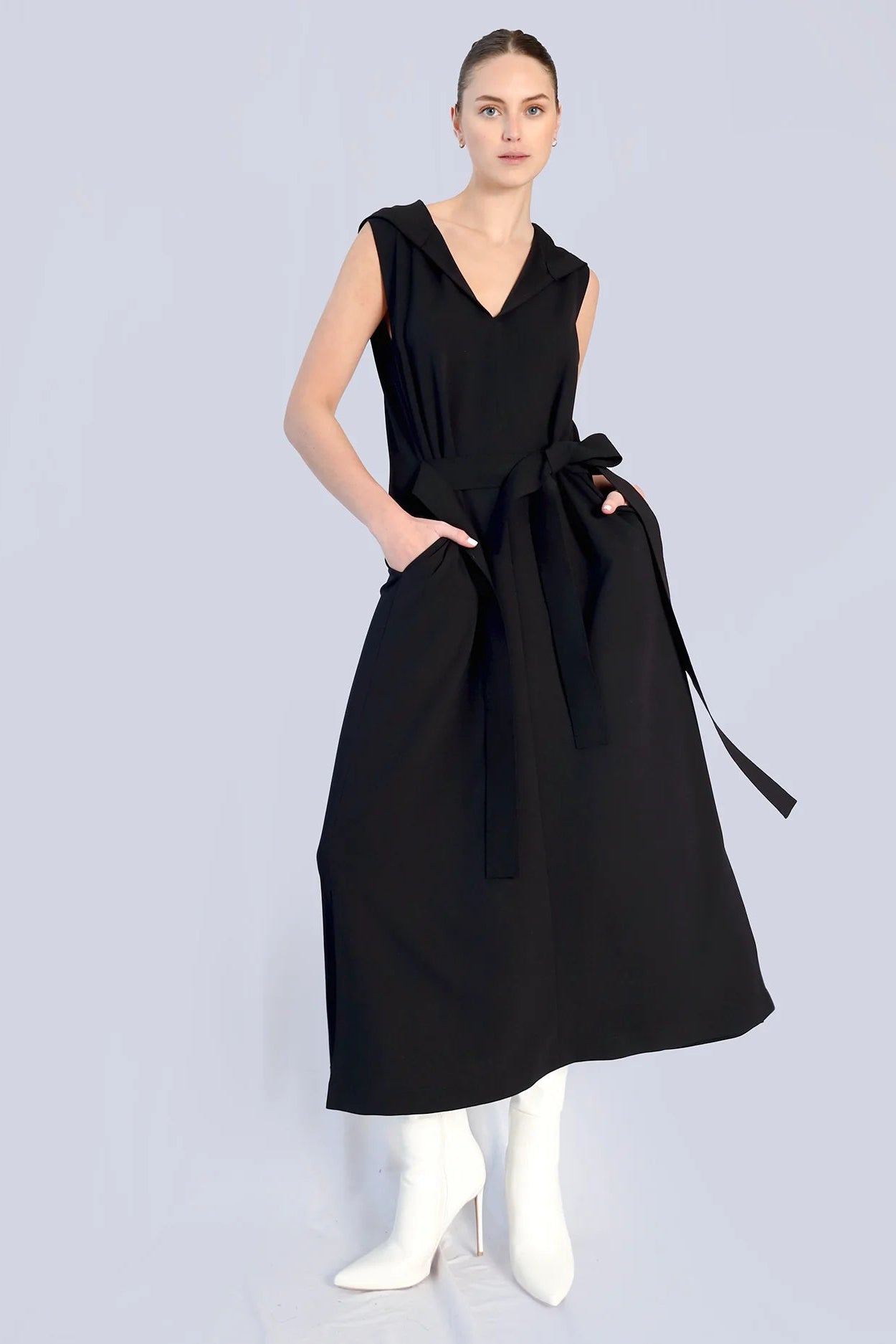 Woman wearing a long black dress with pockets and a tie front with white heeled boots on a grey background.