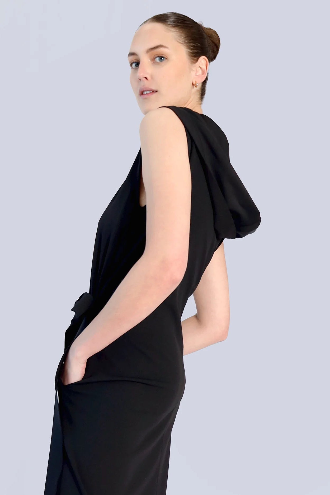 The side of a woman wearing a black dress with pockets and a hood on a grey background.