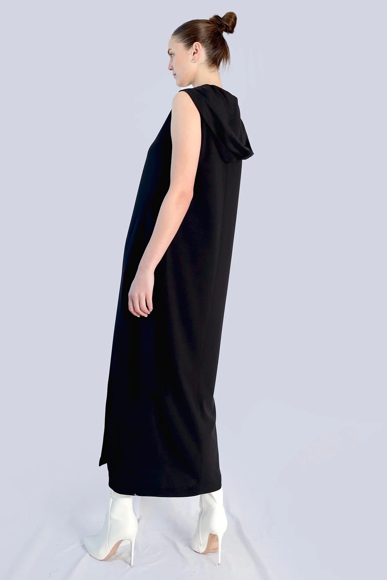 The back/side angle of a woman wearing a long black dress with a hood with white heeled boots on a grey background.
