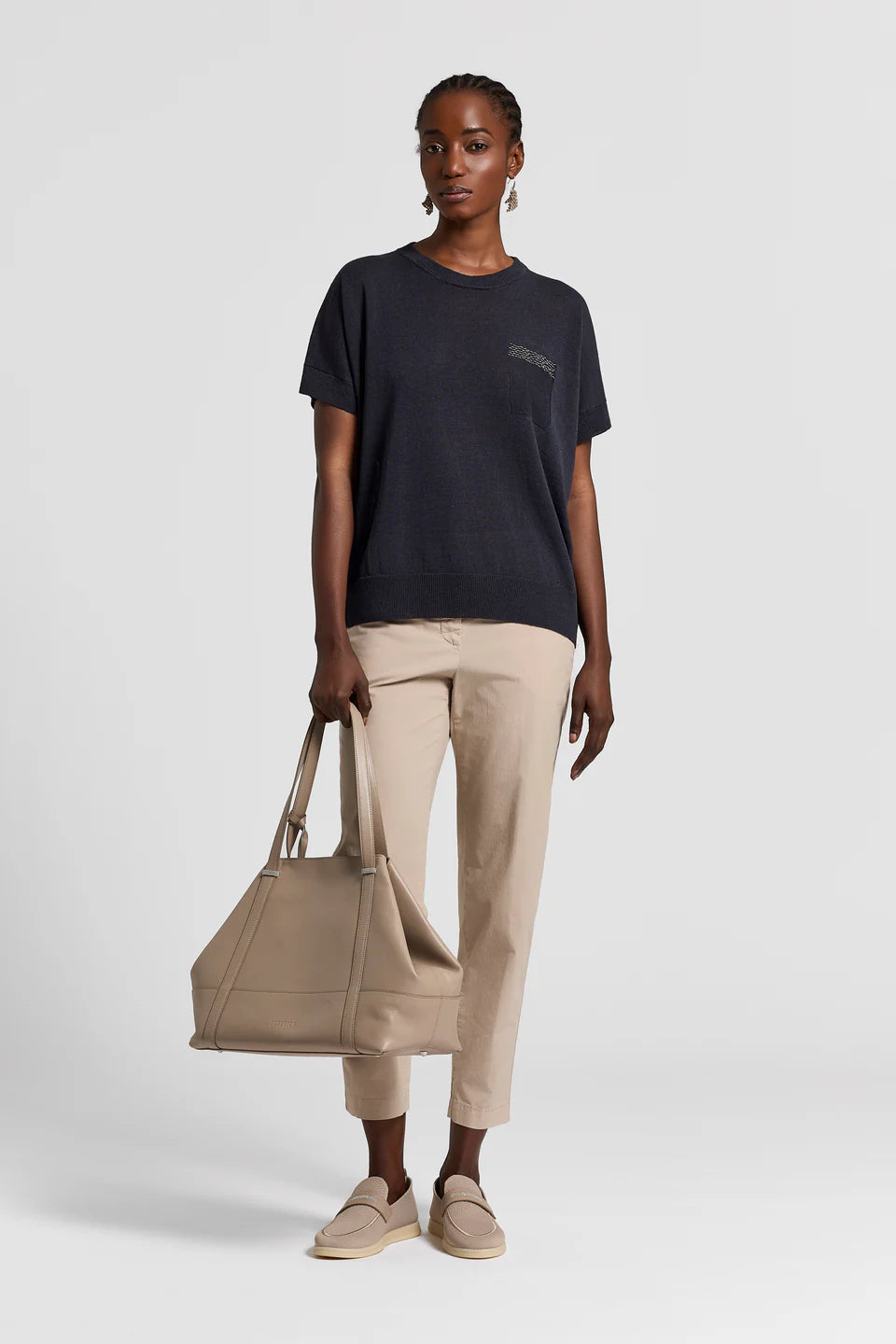 Woman holding a beige bag wearing a navy t-shirt and beige pants on a white background.