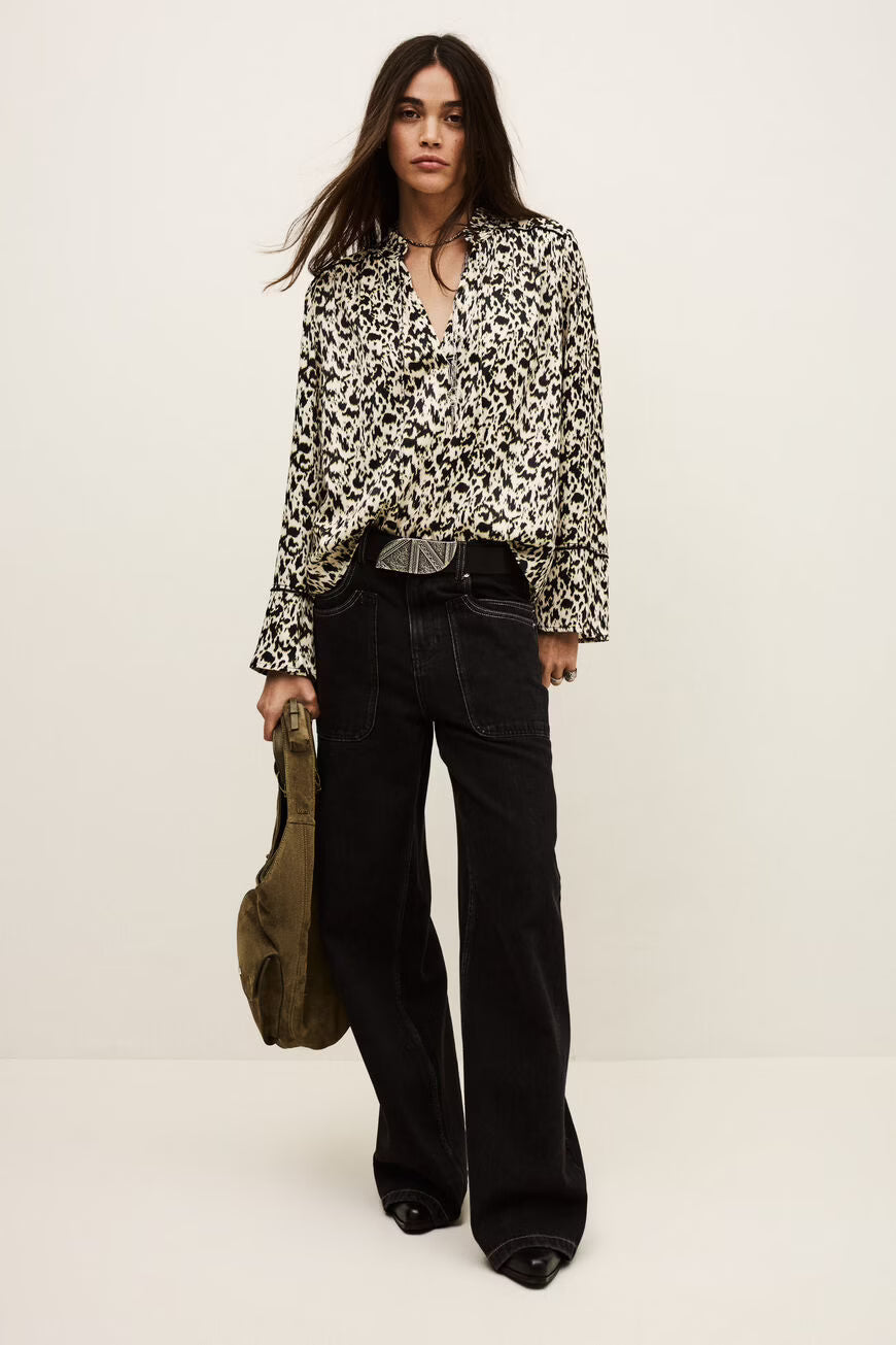 Woman wearing a patterned blouse and black pants holding a green bag on a white background.
