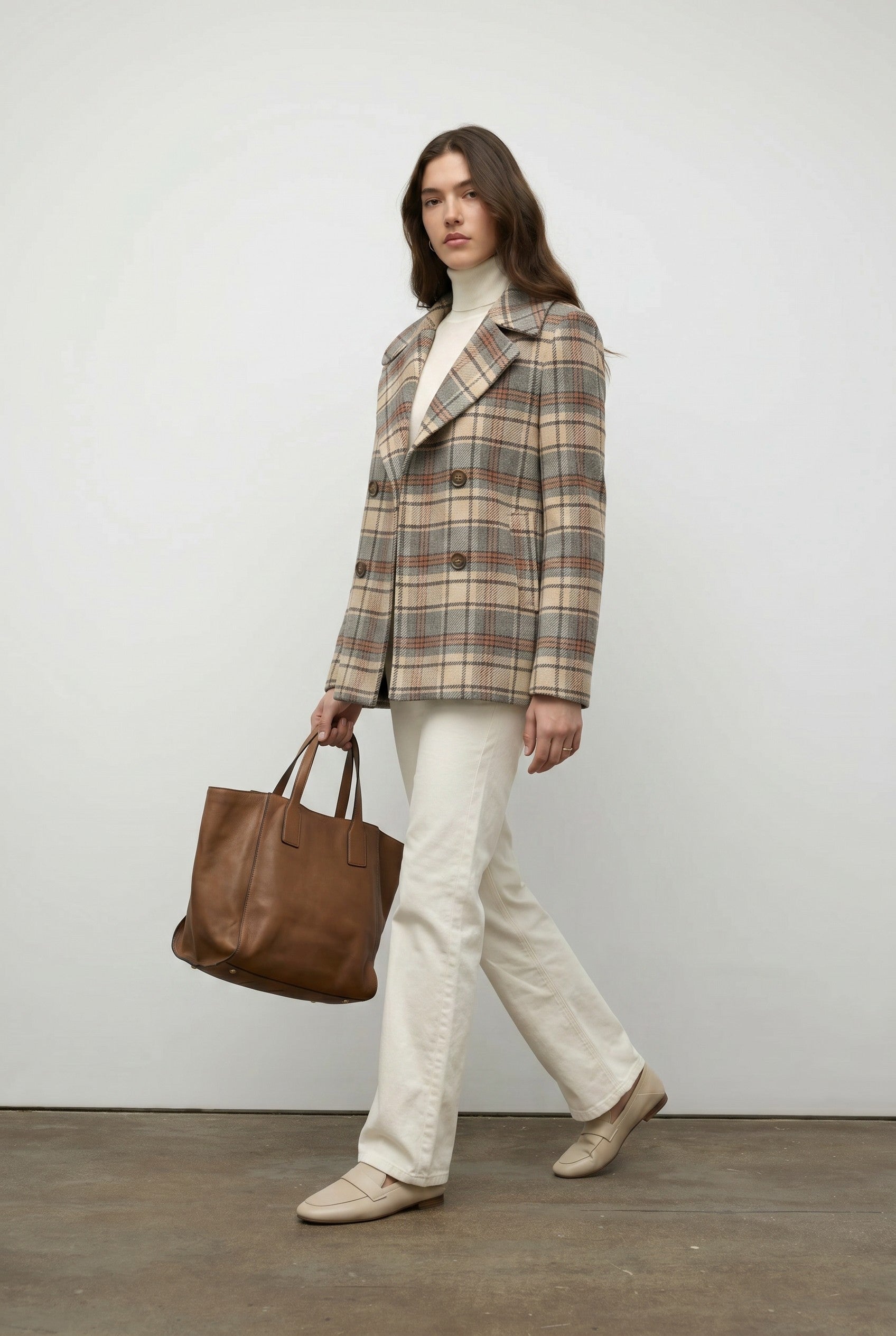 Woman wearing a plaid coat and holding a brown bag against a plain background.