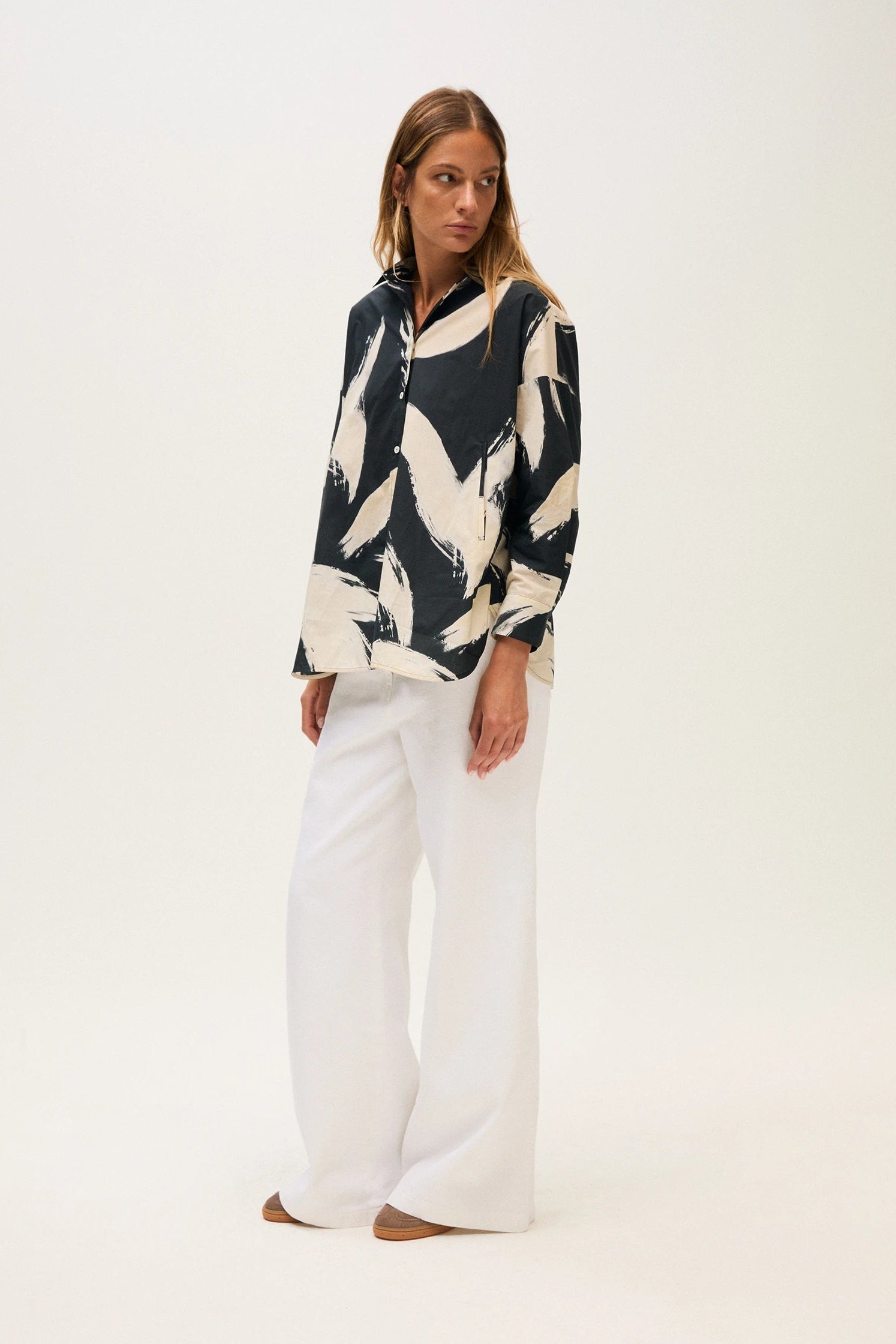 Woman wearing a patterned top and white pants on a white background.