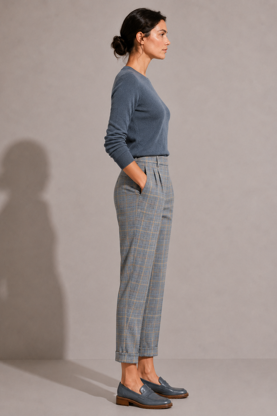 Side view of model wearing the blue-gray plaid Maura Pant, highlighting the tailored fit, front pleats, side pocket, and cuffed hem.