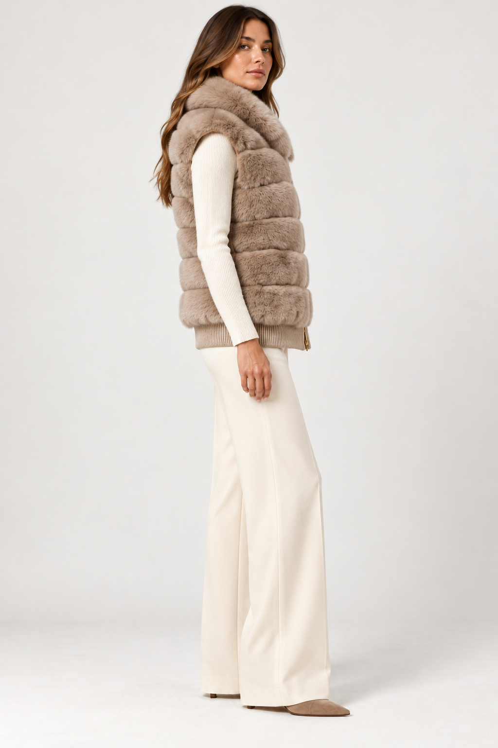 Side view of model wearing the Suprema Rex rabbit fur vest in soft taupe, featuring plush horizontal fur panels, a voluminous shawl collar, sleeveless silhouette, and ribbed wool-cashmere hem. Styled over an ivory knit top with cream wide-leg trousers and taupe pointed-toe shoes against a neutral studio background.