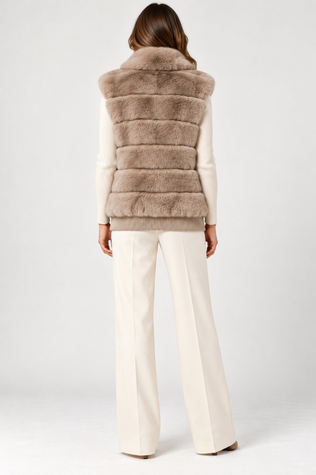 Back view of model wearing the Suprema Rex rabbit fur vest in soft taupe, featuring plush horizontal fur panels, a voluminous fur collar, sleeveless silhouette, and ribbed wool-cashmere hem. Styled over an ivory knit top with cream wide-leg trousers against a neutral studio background.