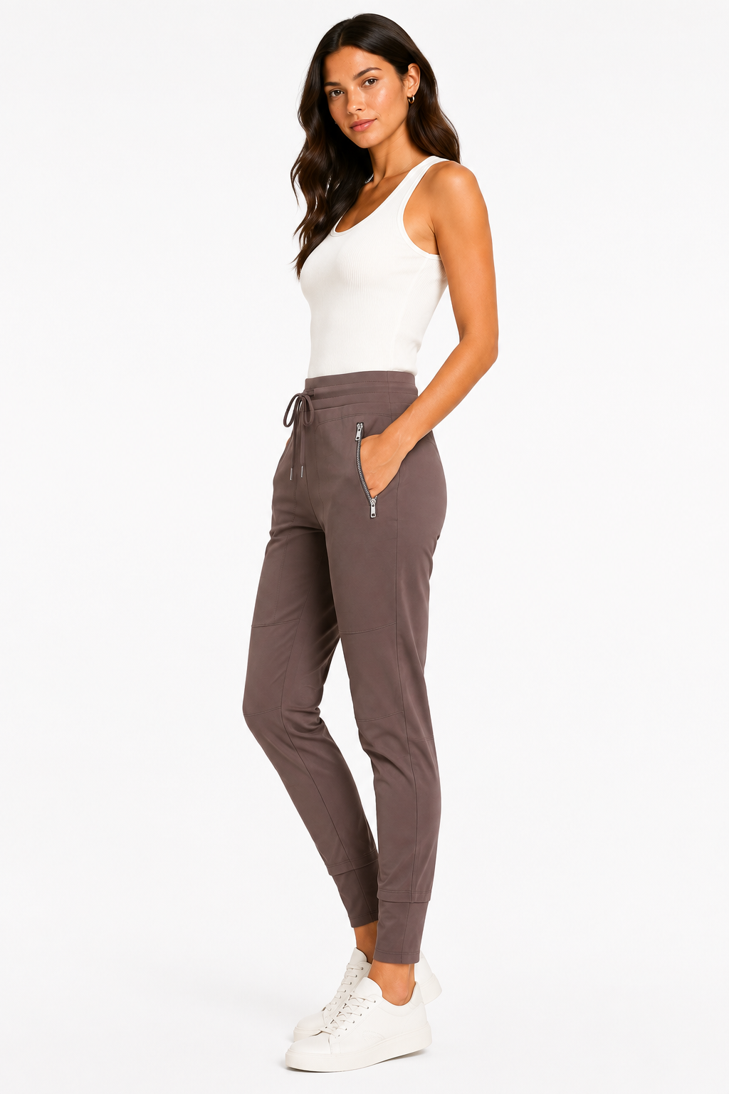 A woman modeling a pair of high-waisted, ankle-length jogger pants in a muted taupe-brown color. The pants feature a drawstring waist, zippered side pockets, and elastic cuffs.
