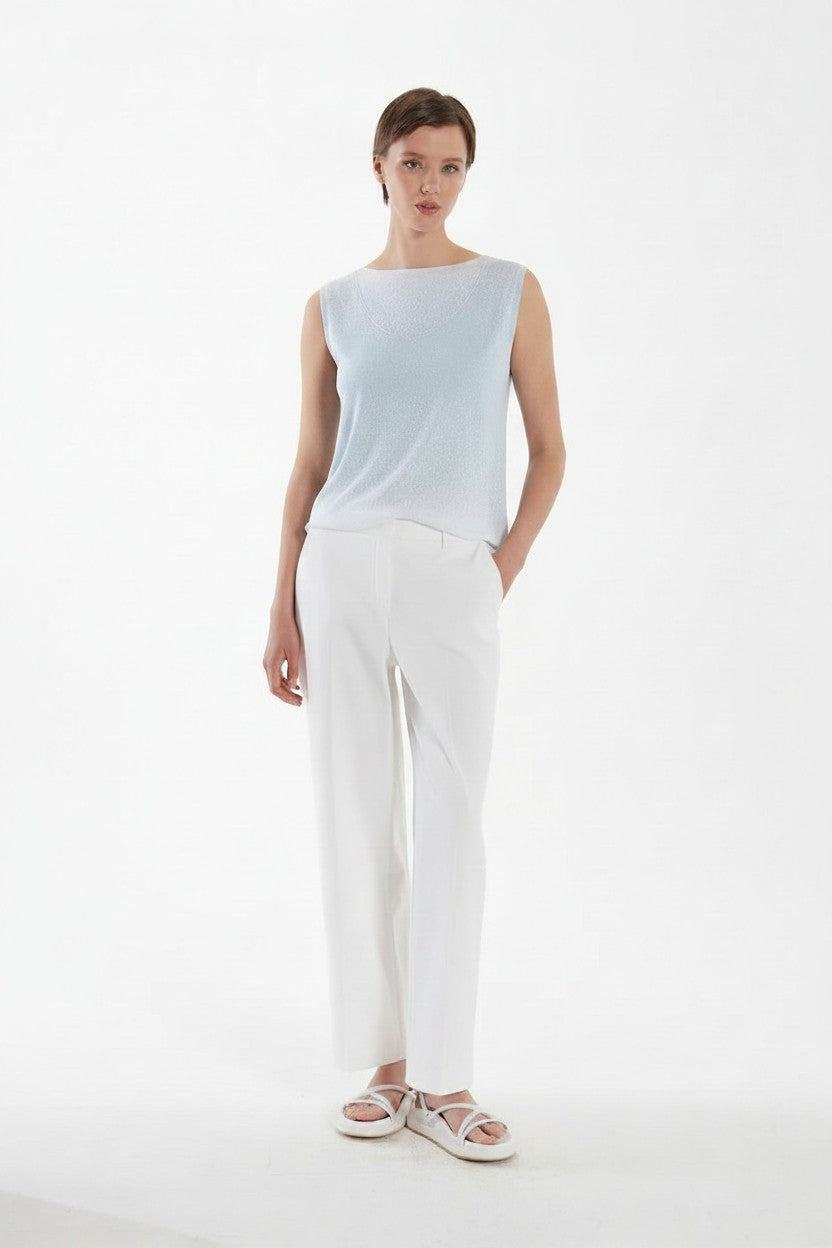 Woman wearing a light blue sleeveless top and white pants on a white background.