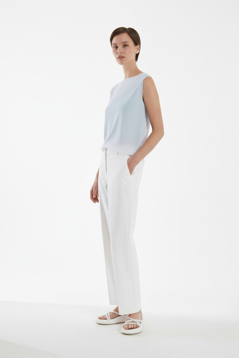 Woman wearing a light blue sleeveless top and white pants on a white background.
