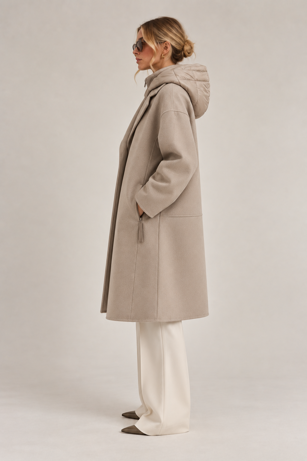 Side view of model wearing a taupe long coat with quilted hood, relaxed sleeves, and ivory wide-leg trousers.