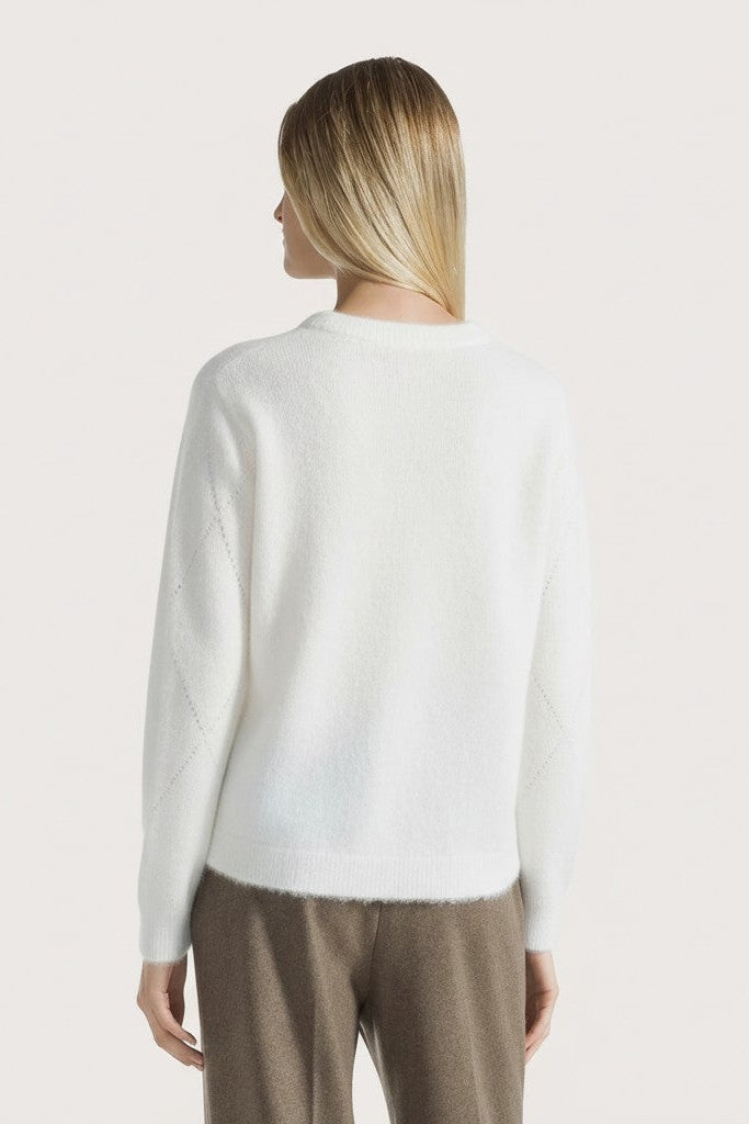 Woman wearing a white sweater and brown pants on a light gray background.