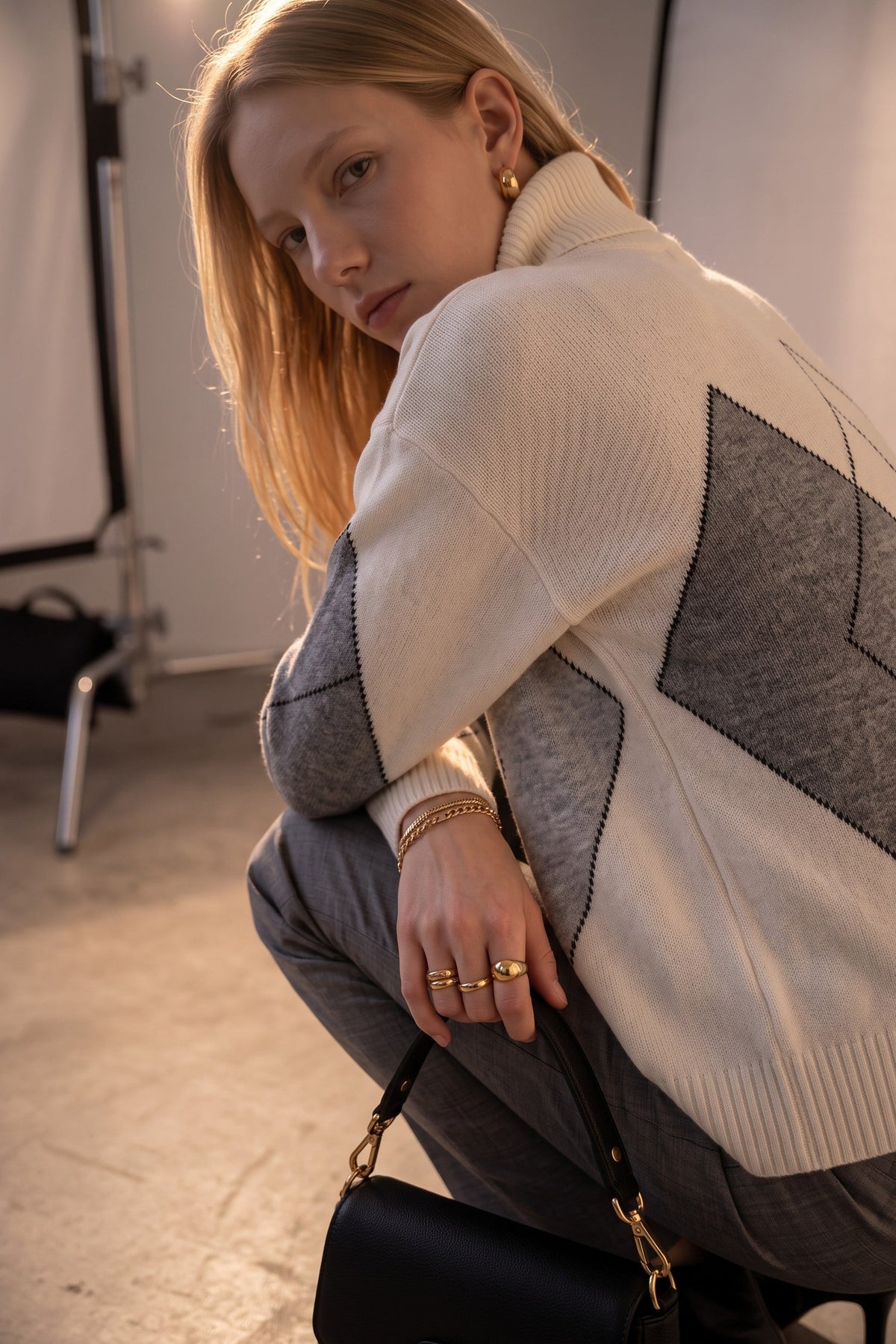 A woman crouches in a studio setting wearing a cream and gray geometric patterned knit sweater, gray trousers, and gold jewelry, holding a black handbag.