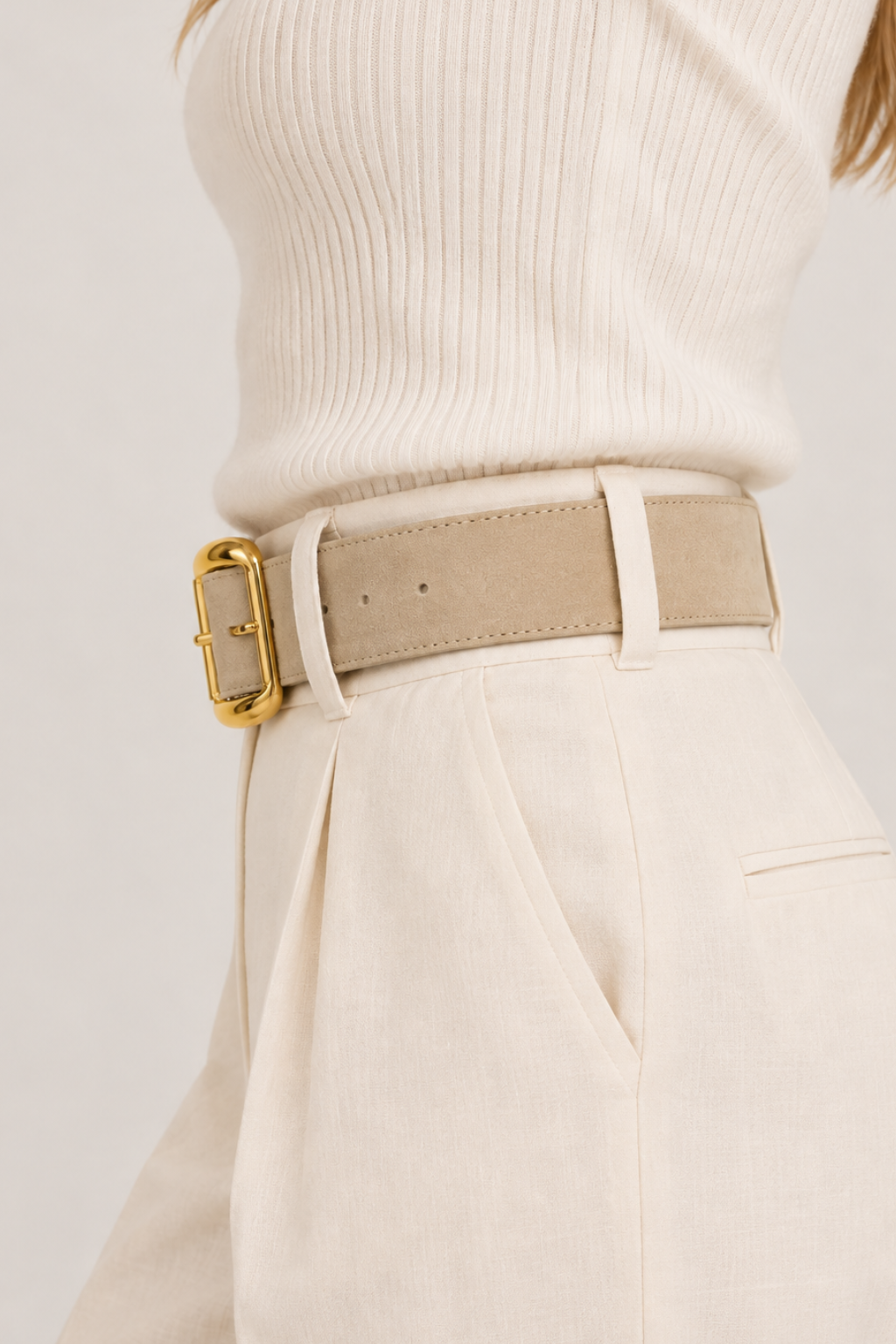 A side view close-up of a model wearing a beige wide belt with a prominent gold buckle, styled with cream-colored trousers and a ribbed cream top against a light off-white background.