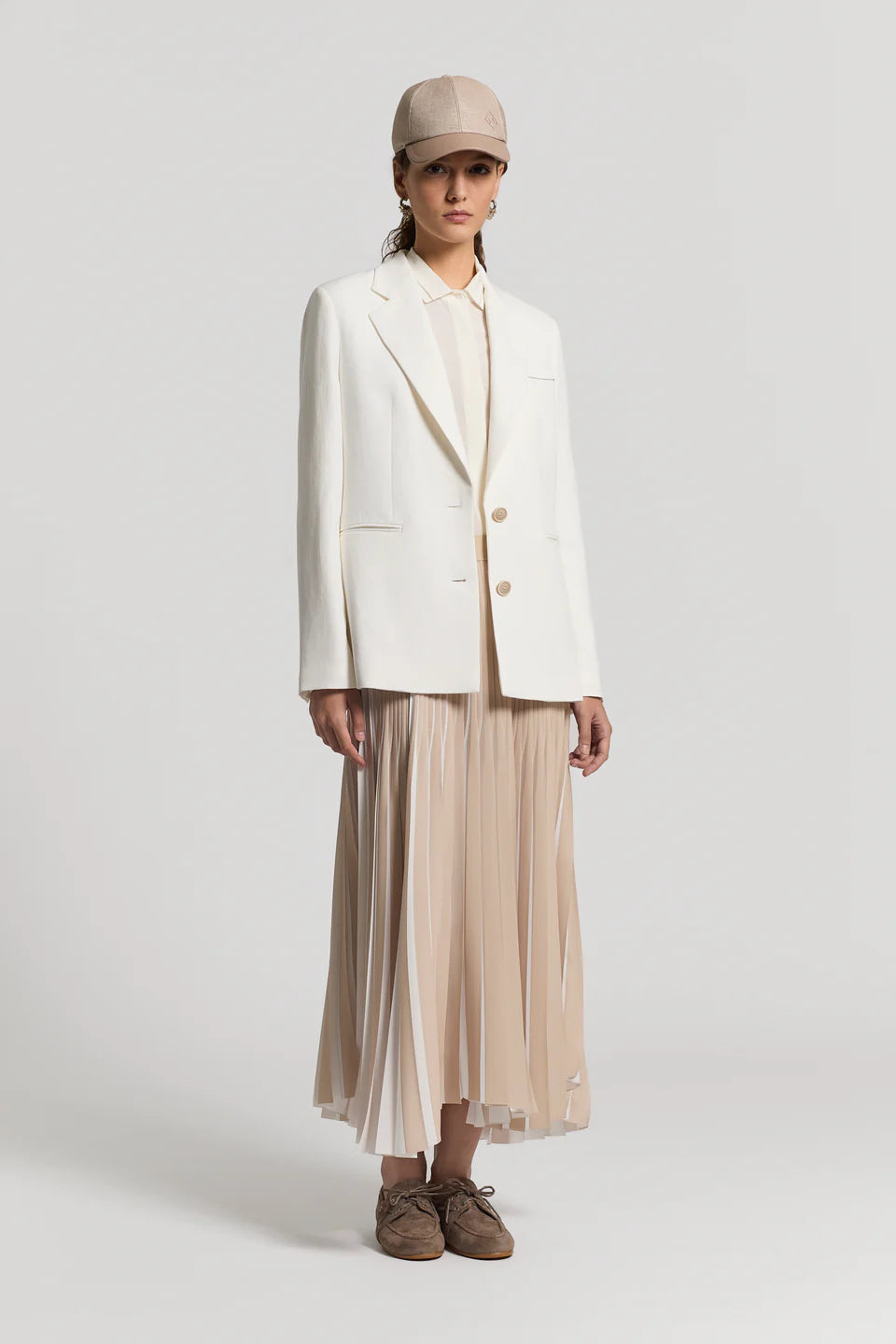 Woman wearing a white blouse underneath a white blazer, with a beige skirt, and a hat on a light gray background.