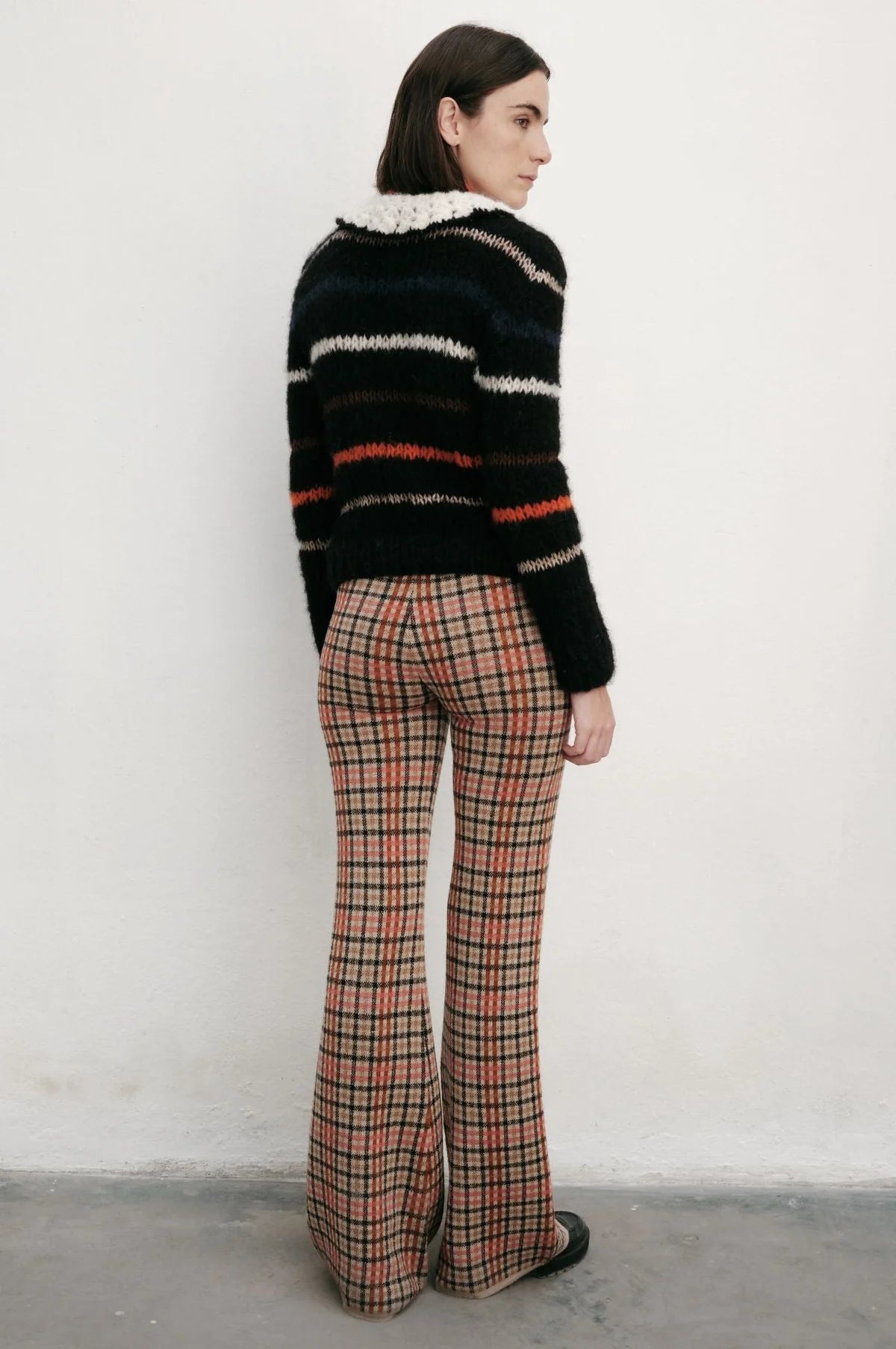 Woman wearing a striped sweater and plaid pants against a white wall.