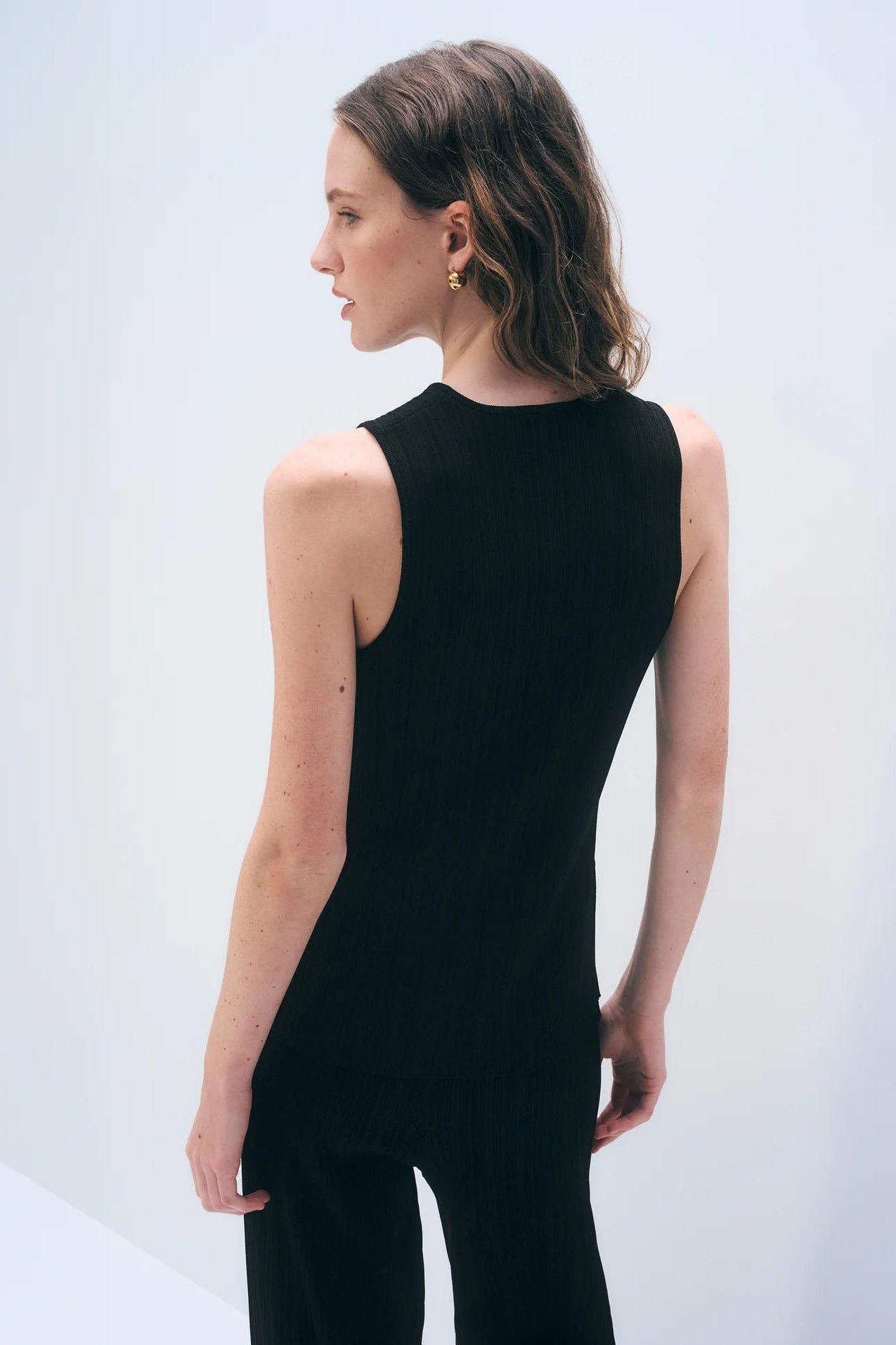 The back of a woman wearing a black sleeveless top and pants against a light gray background.