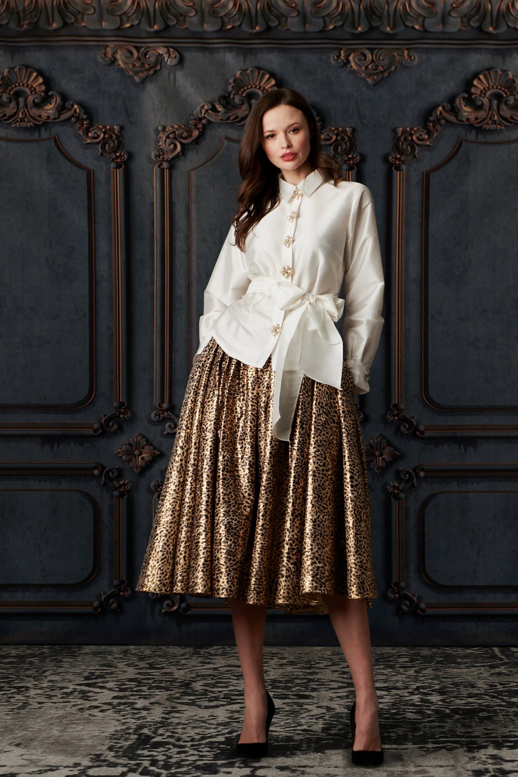 Woman wearing a white blouse and gold patterned skirt against an ornate wall.