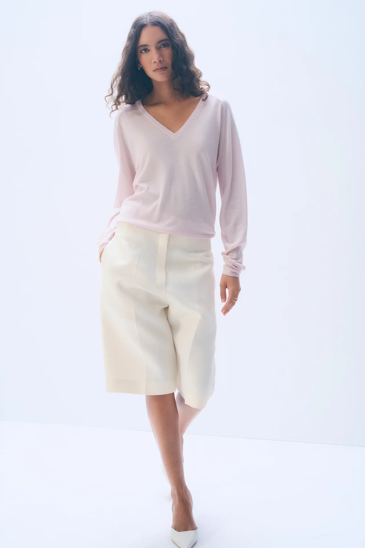 Woman wearing a light pink sweater and white pants on a white background.