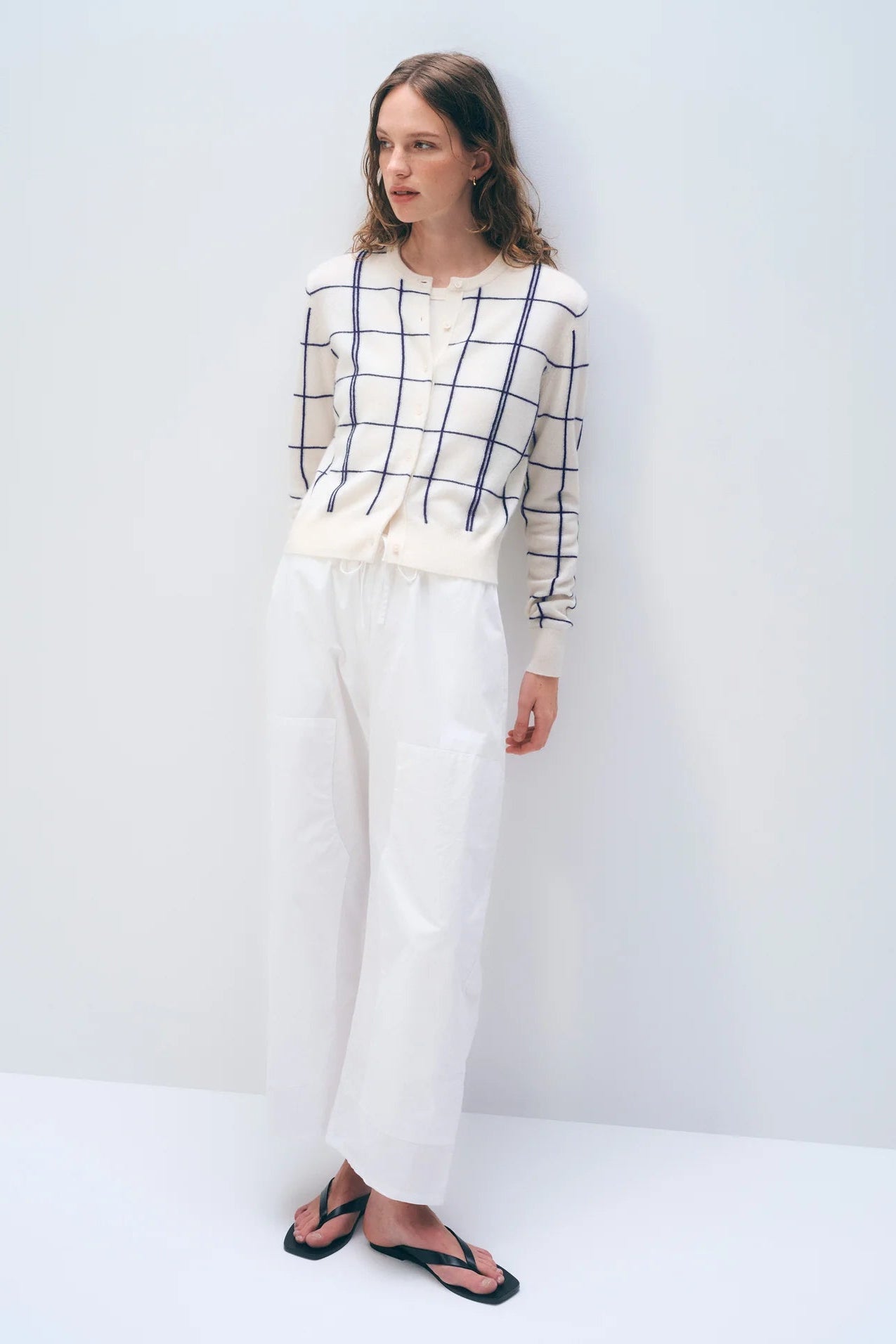 Woman wearing a checkered cardigan and white pants against a plain background.