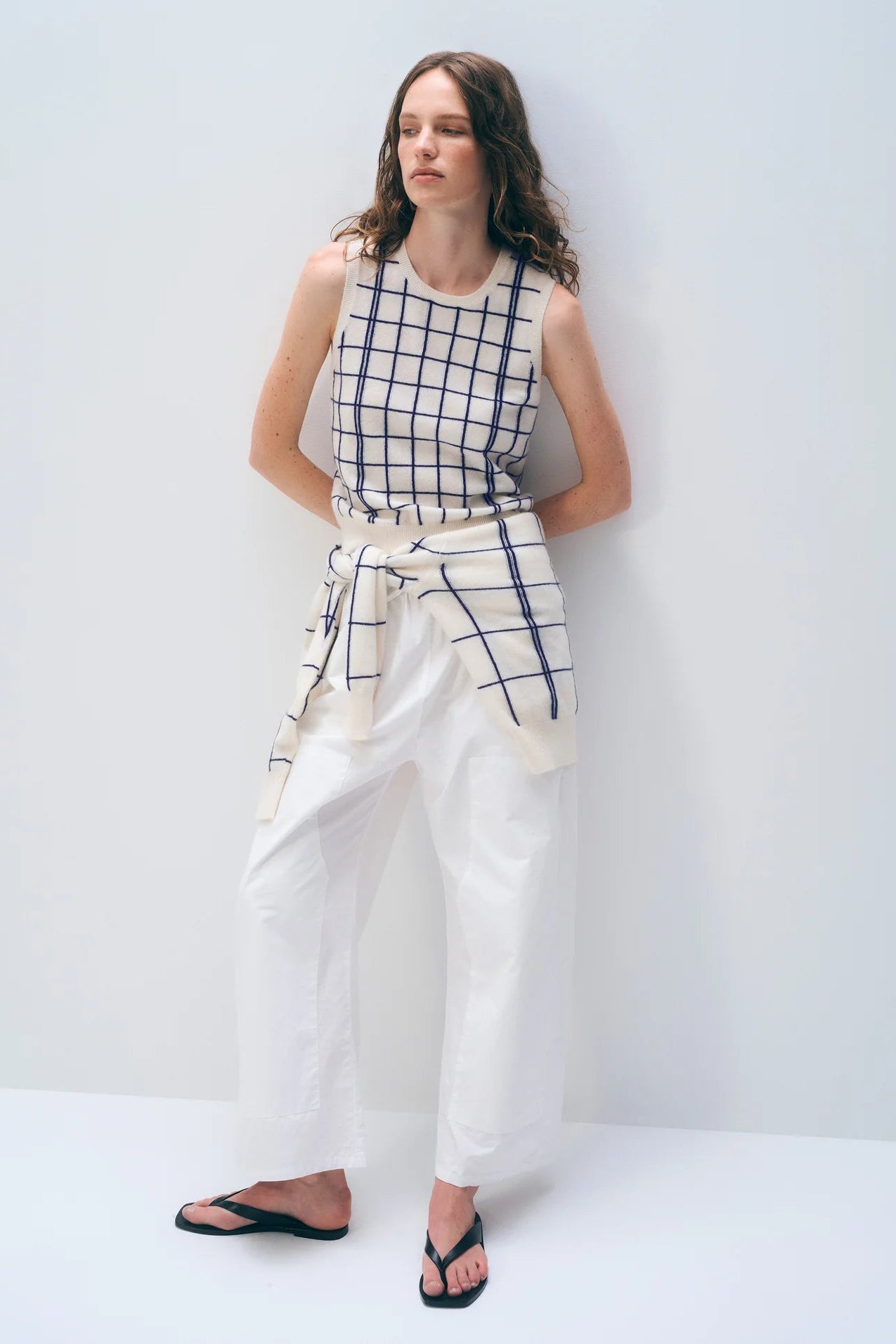 Woman wearing a sleeveless checkered top and white pants against a plain background.