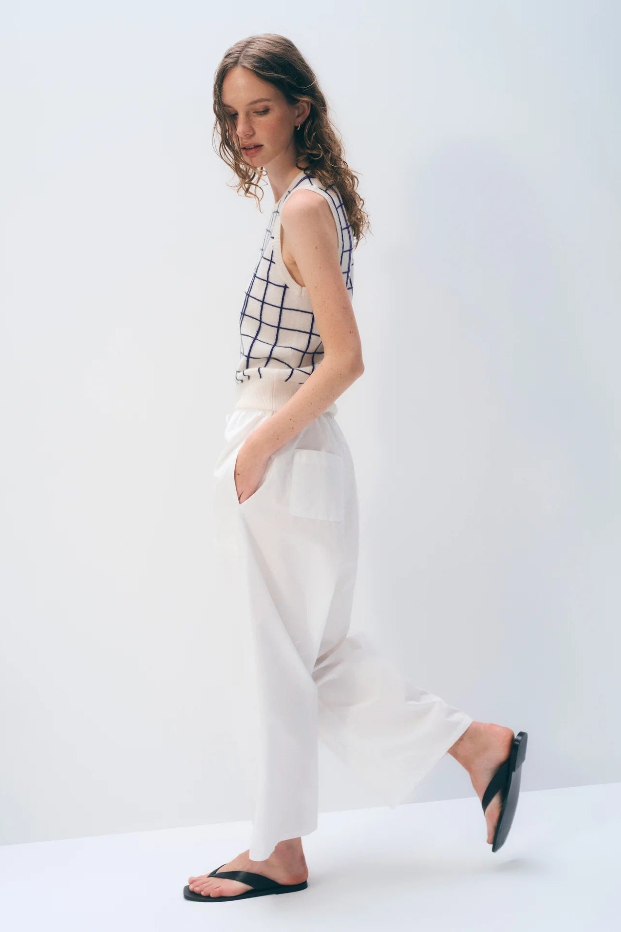 Woman wearing a sleeveless checkered top and white pants on a plain background.