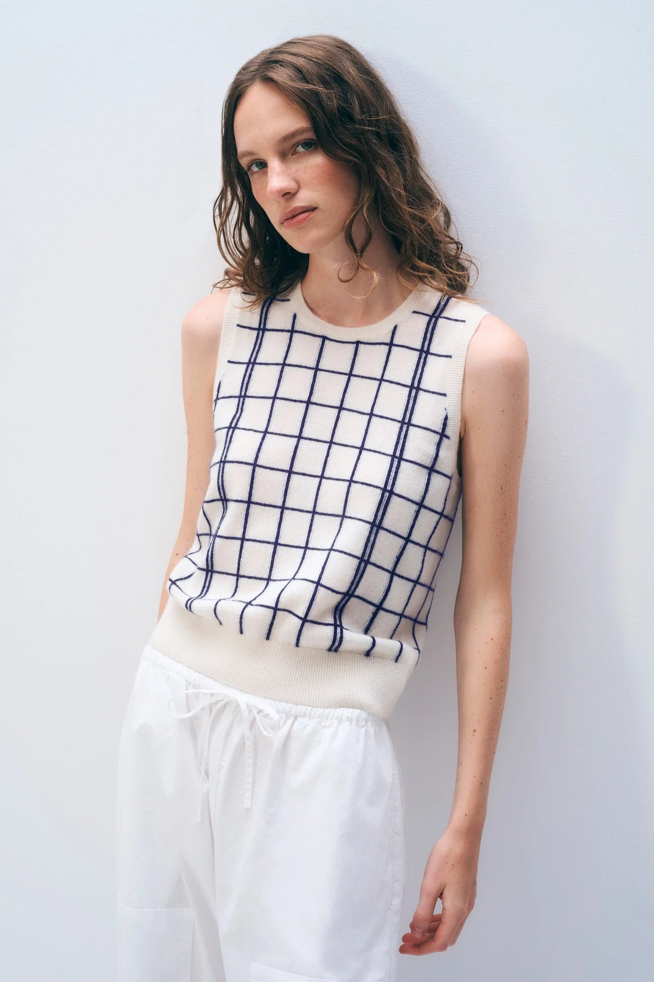Woman wearing a sleeveless checkered top and white pants against a light background.