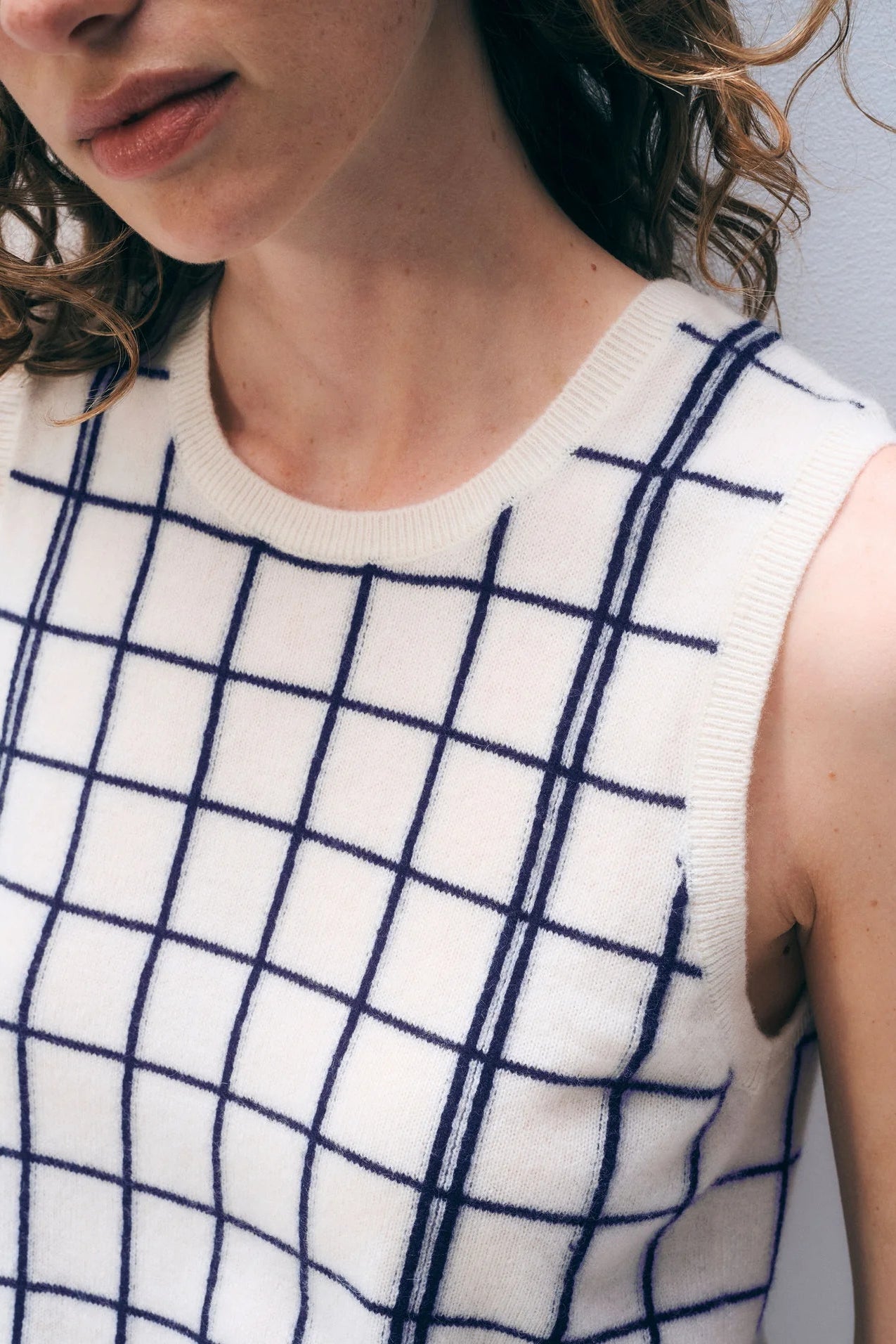Close up of a woman wearing a sleeveless sweater with blue grid pattern on a neutral background.