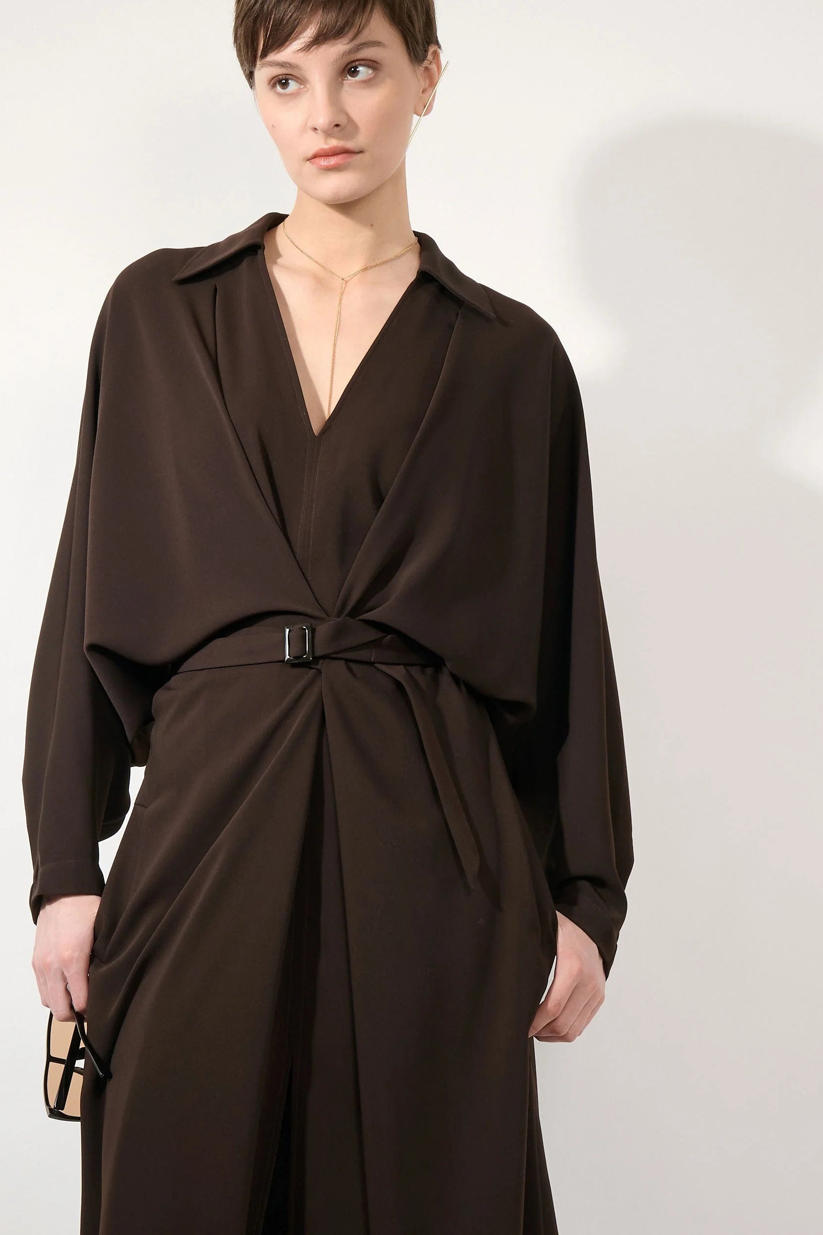 A model wearing a dark brown wrap-style dress with a tie waist and long draped sleeves, photographed against a plain light gray studio background.