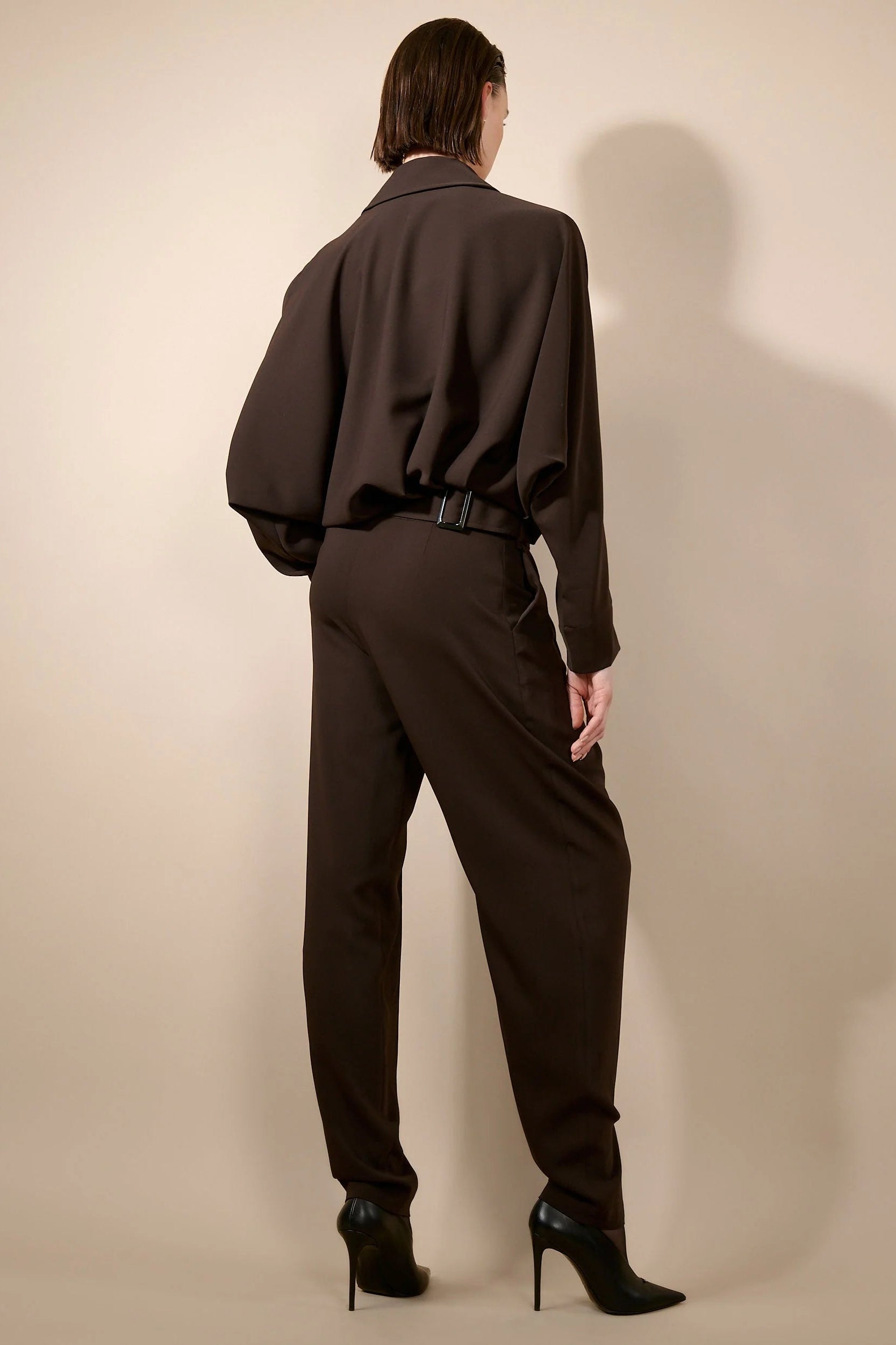 A model shown from the back wearing a dark brown draped long-sleeve top and matching dark brown trousers, paired with black pointed-toe stiletto heels, against a warm beige studio background.