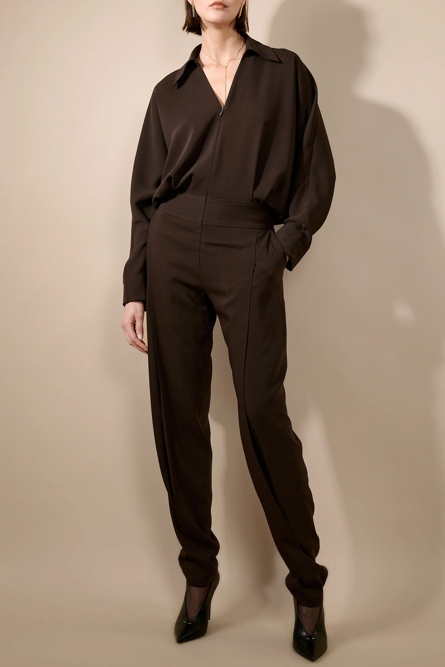 A model wearing a dark brown long-sleeve collared jumpsuit with a high-waisted, relaxed fit, paired with black pointed-toe heels, set against a plain beige studio background.