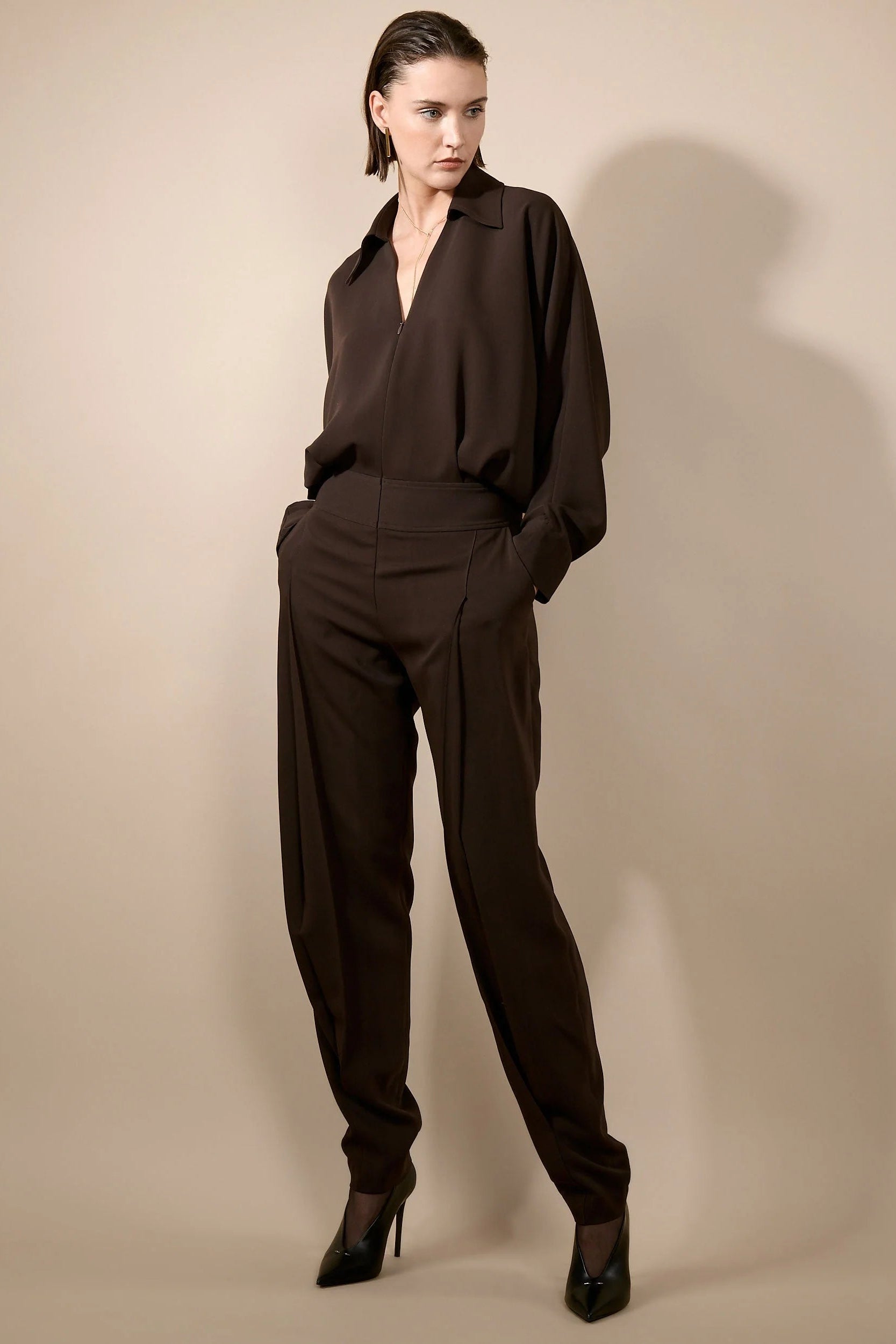 A model wearing a dark brown long-sleeve collared jumpsuit with matching trousers and black high heels, standing against a plain beige studio background.