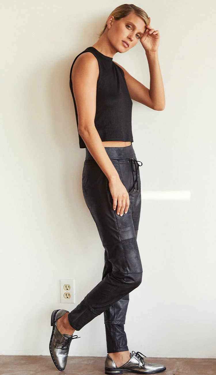 Woman wearing a black sleeveless top and dark pants against a white wall.