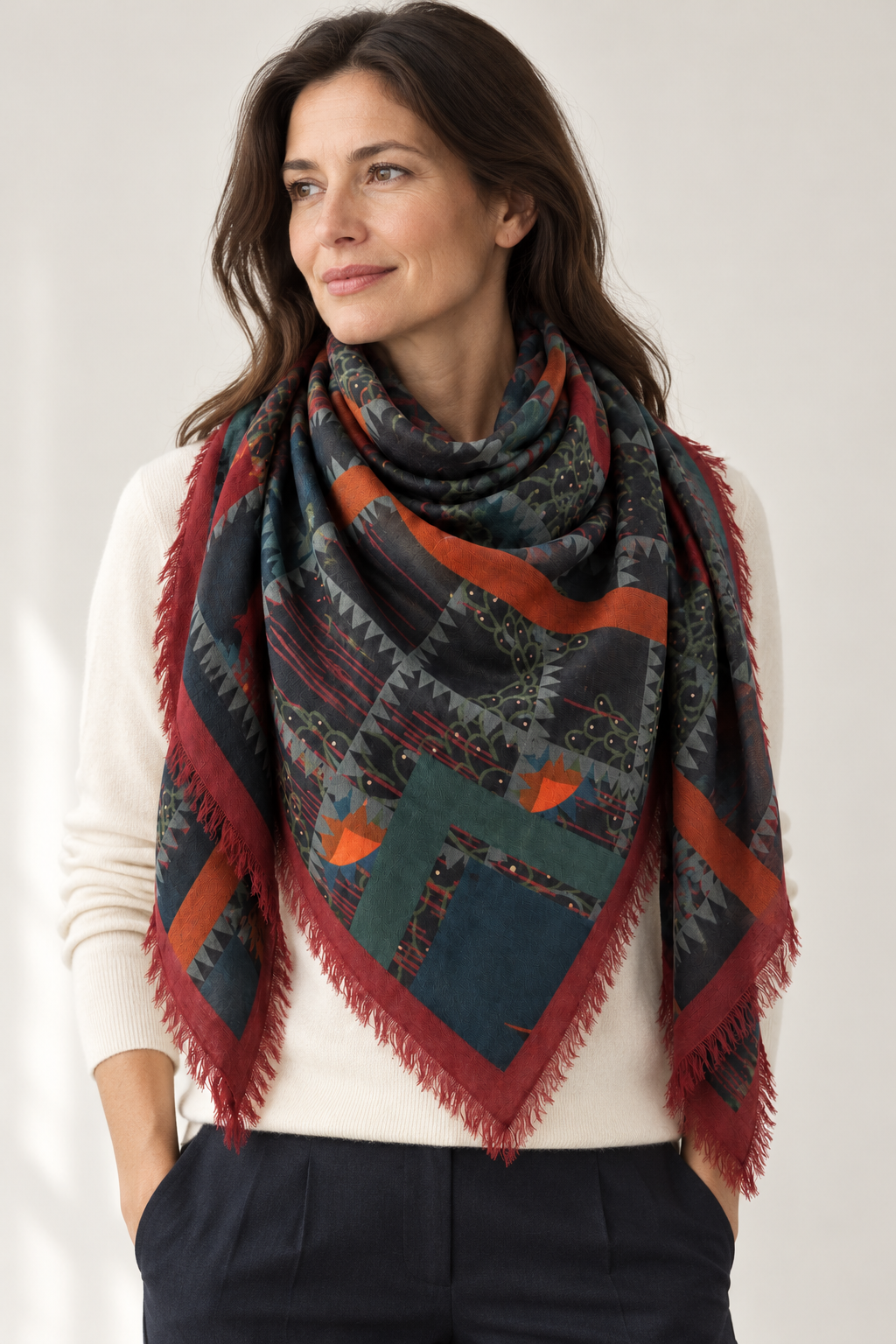 Woman wearing the Franco Ferrari Magar botanical geometric wool scarf with teal and rust accents and burgundy fringe over an ivory sweater.