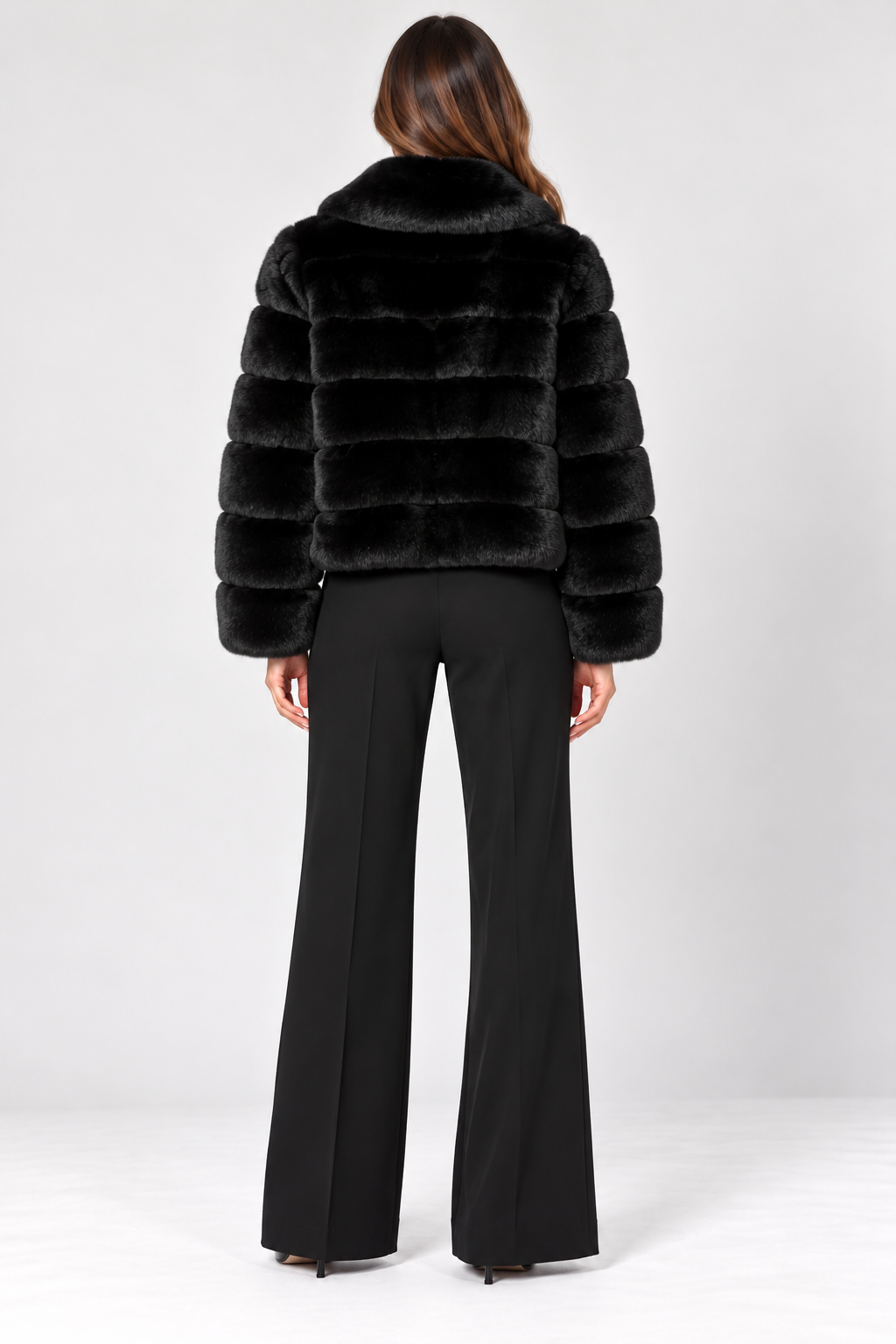 Back view of model wearing the Suprema Black Rex Rabbit Fur Bomber Jacket, featuring luxurious genuine Rex rabbit fur, horizontal fur panels, an oversized fold-down collar, long sleeves, and a relaxed hip-length silhouette. Styled with tailored black wide-leg trousers and pointed-toe heels against a neutral studio background.