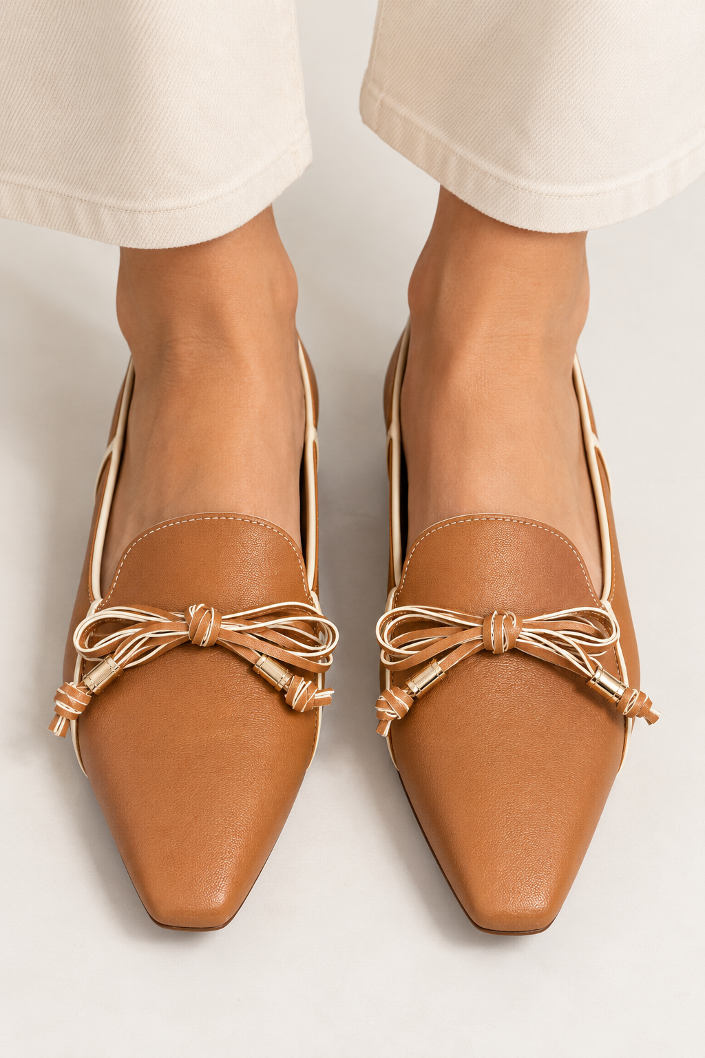 Front close-up of model wearing RODO Antilope camel leather loafers with elongated square toes, ivory contrast piping, delicate two-tone leather bows, and gold-tone accents, styled with cream trousers.
