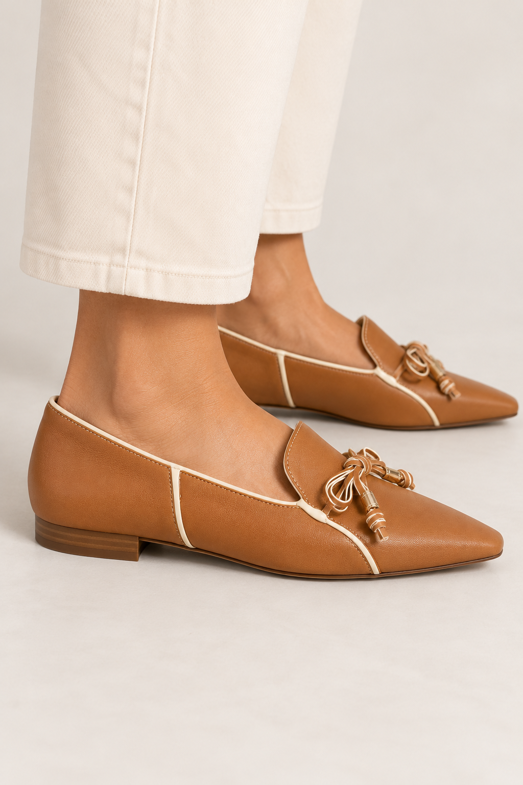 Side-view close-up of model wearing RODO Antilope camel leather loafers featuring ivory contrast piping, delicate two-tone leather bows, gold-tone accents, elongated square toes, and low stacked heels, styled with cropped cream trousers.