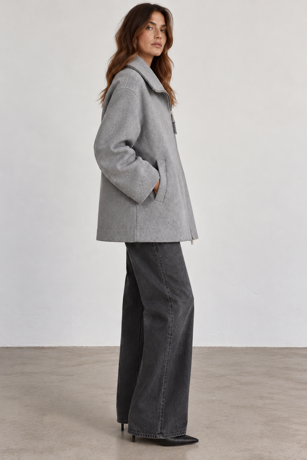 Side view of model wearing a Seventy gray wool-blend coat without the removable padded collar, featuring a relaxed, boxy silhouette, structured fold-down collar, long sleeves, and side pockets, styled with charcoal wide-leg jeans and black pointed-toe boots.